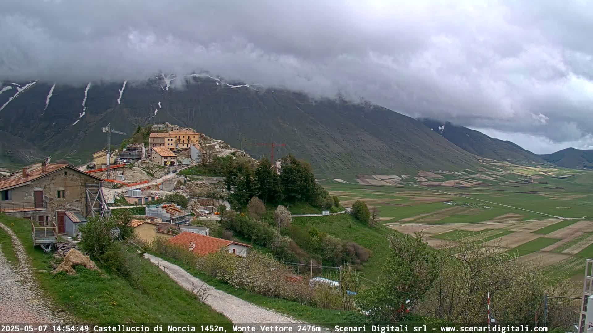 A partly cloudy day reveals a hilly landscape with a village nestled against snow-capped mountains and a valley of patchwork farmland below.