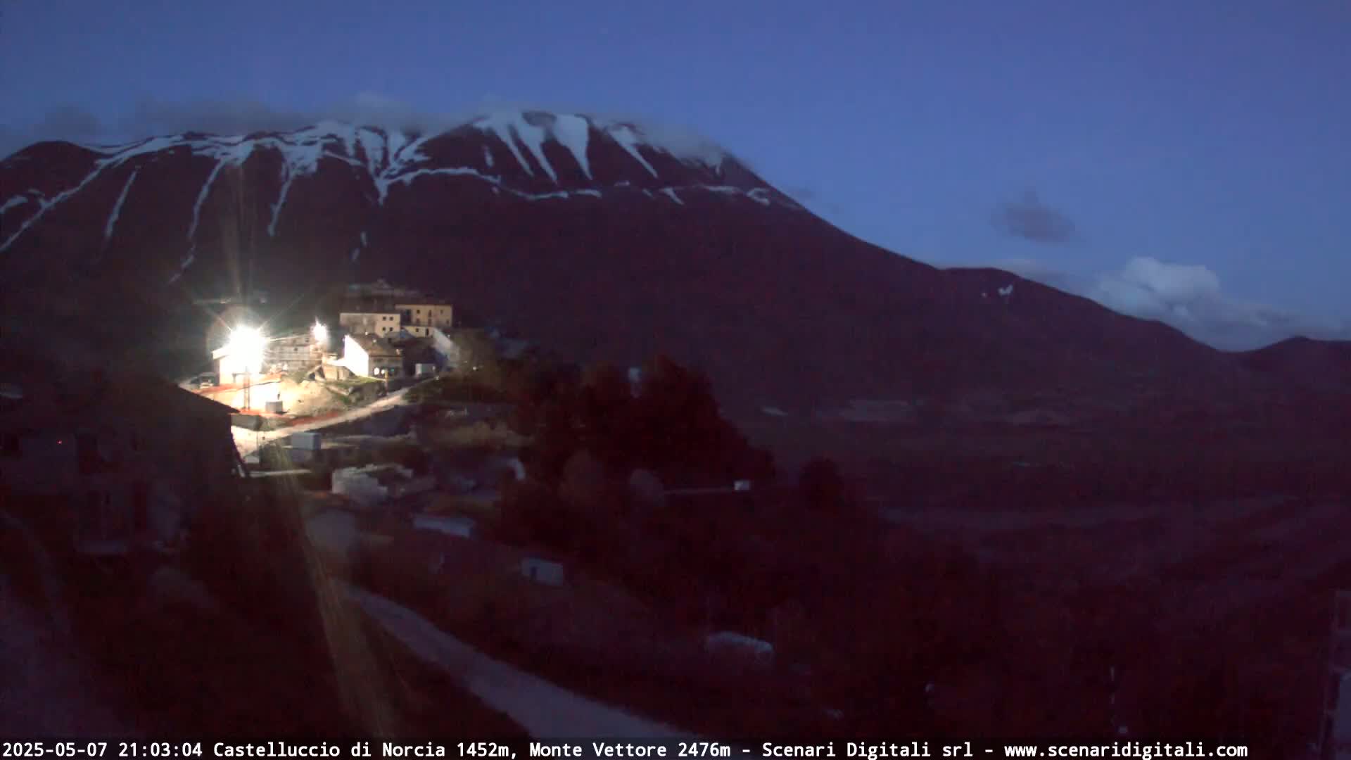 A small village is illuminated at night, nestled below a large, snow-capped mountain under a dark blue twilight sky.