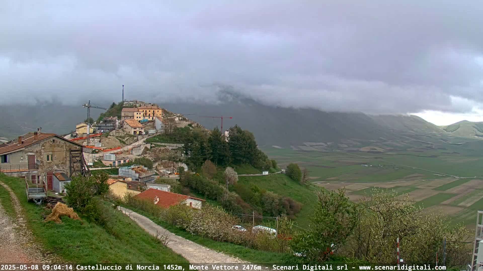A partially cloudy day reveals a hilltop village under construction, overlooking a vast green valley and distant, fog-covered mountains.