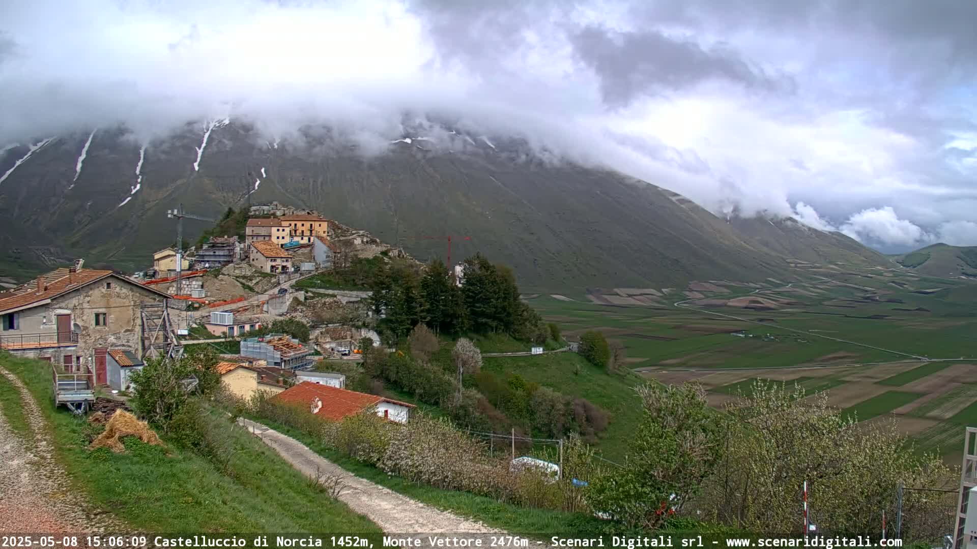 A small village nestled in a valley between green rolling hills and a cloudy, snow-dusted mountain range.