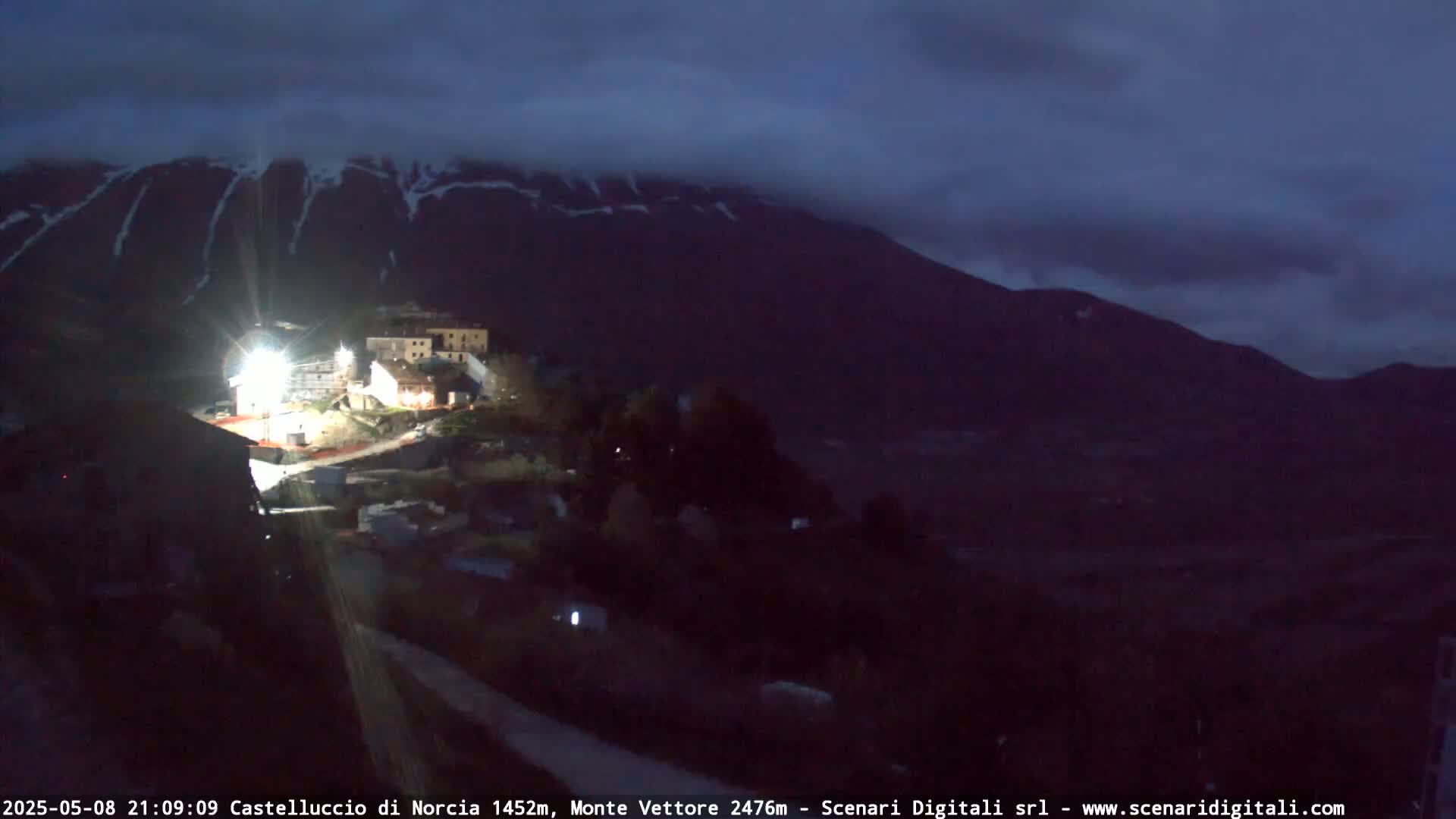 A small village is nestled in a valley beneath a dark, cloudy sky and a snow-capped mountain.
