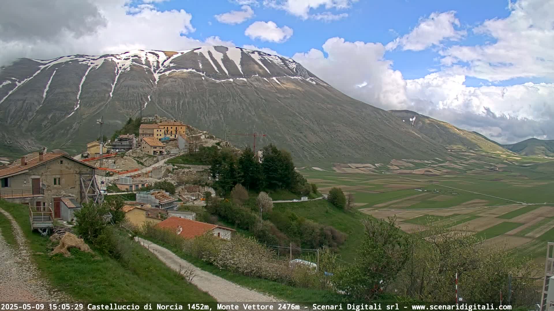 A partly cloudy day reveals a mountain range with snow-capped peaks overlooking a small village nestled in a valley of green fields.
