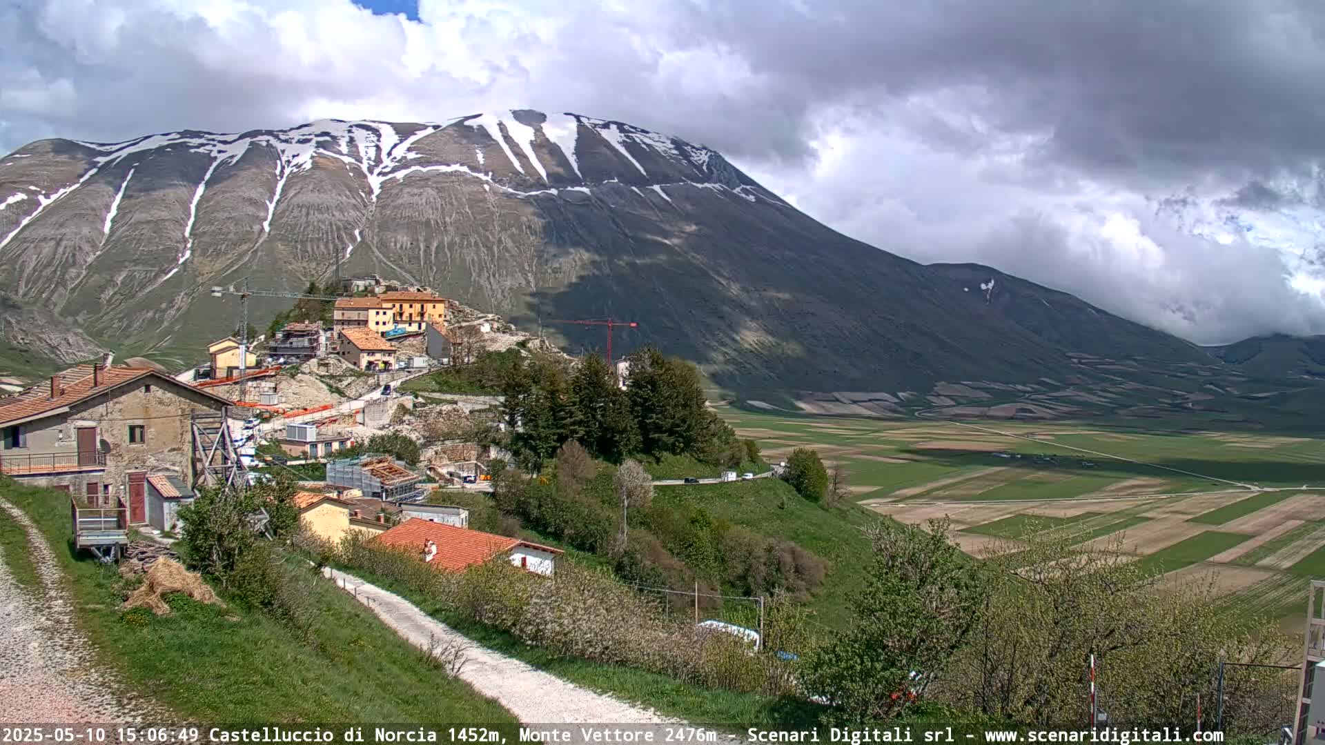 A partially cloudy day reveals a village nestled on a hillside below a snow-dusted mountain range, overlooking a patchwork of green fields.
