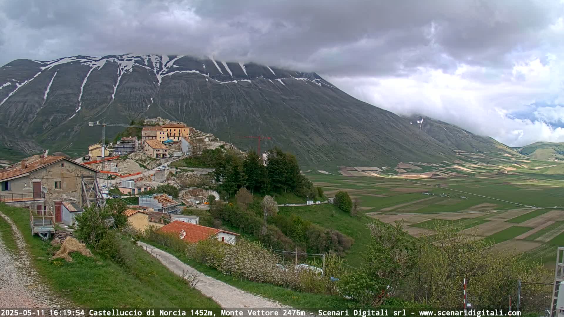 A partially cloudy day reveals a village nestled on a hillside beneath a snow-dusted mountain range, with green farmland stretching out into the valley.