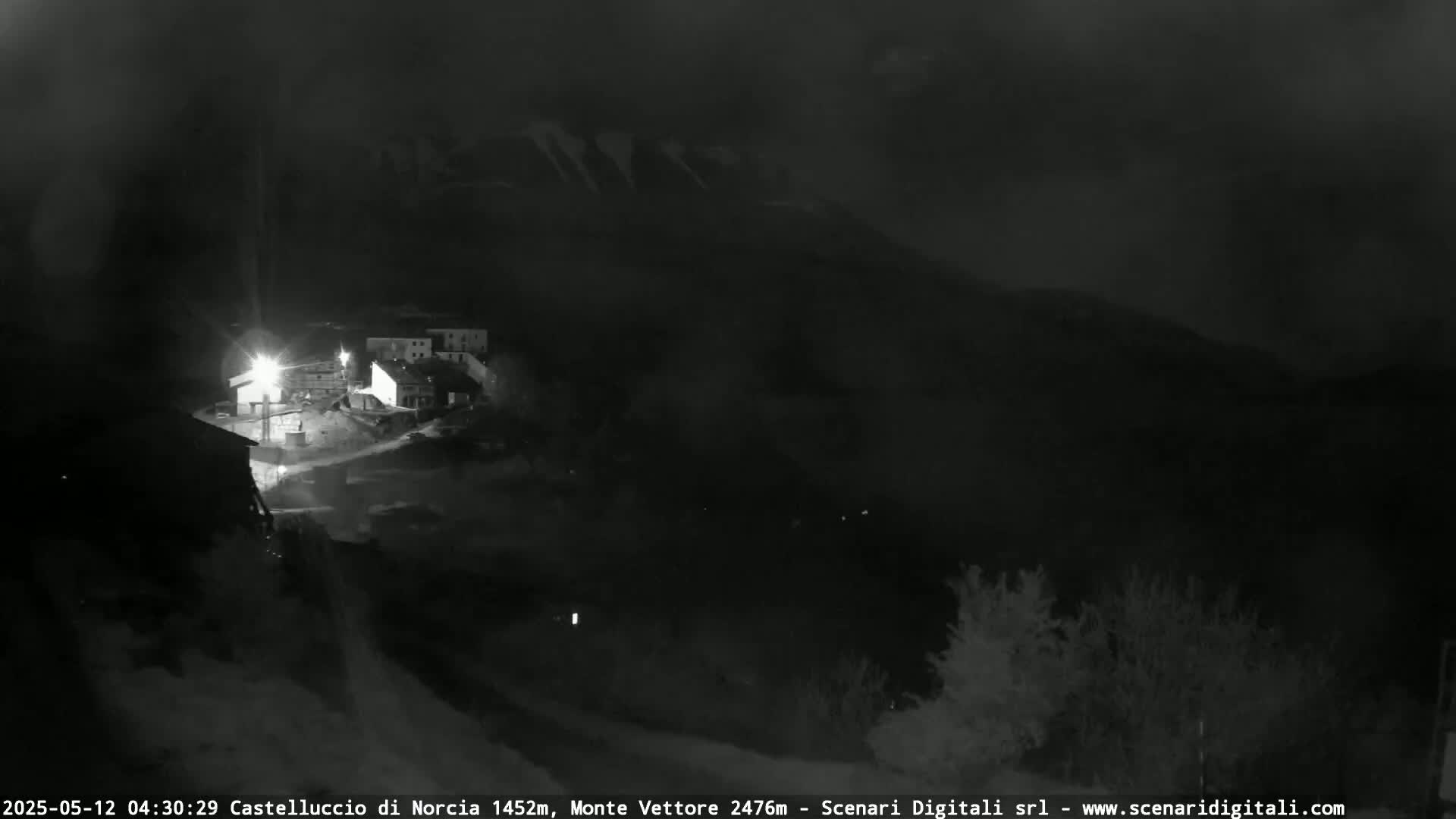 A nighttime, low-light view shows a small village nestled in a valley below dark mountains under a cloudy sky.