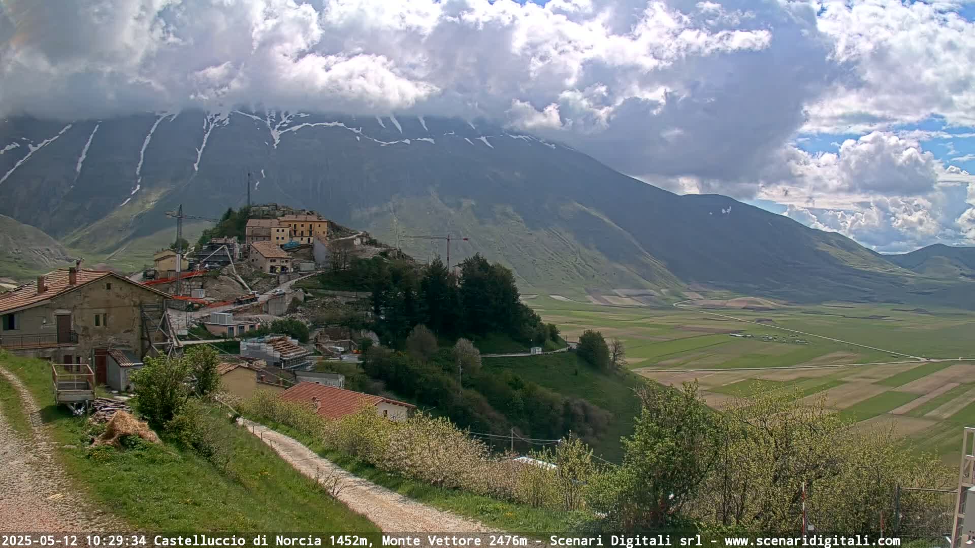 A partly cloudy day reveals a mountain village nestled on a hillside overlooking a vast, verdant valley and snow-capped mountains in the distance.