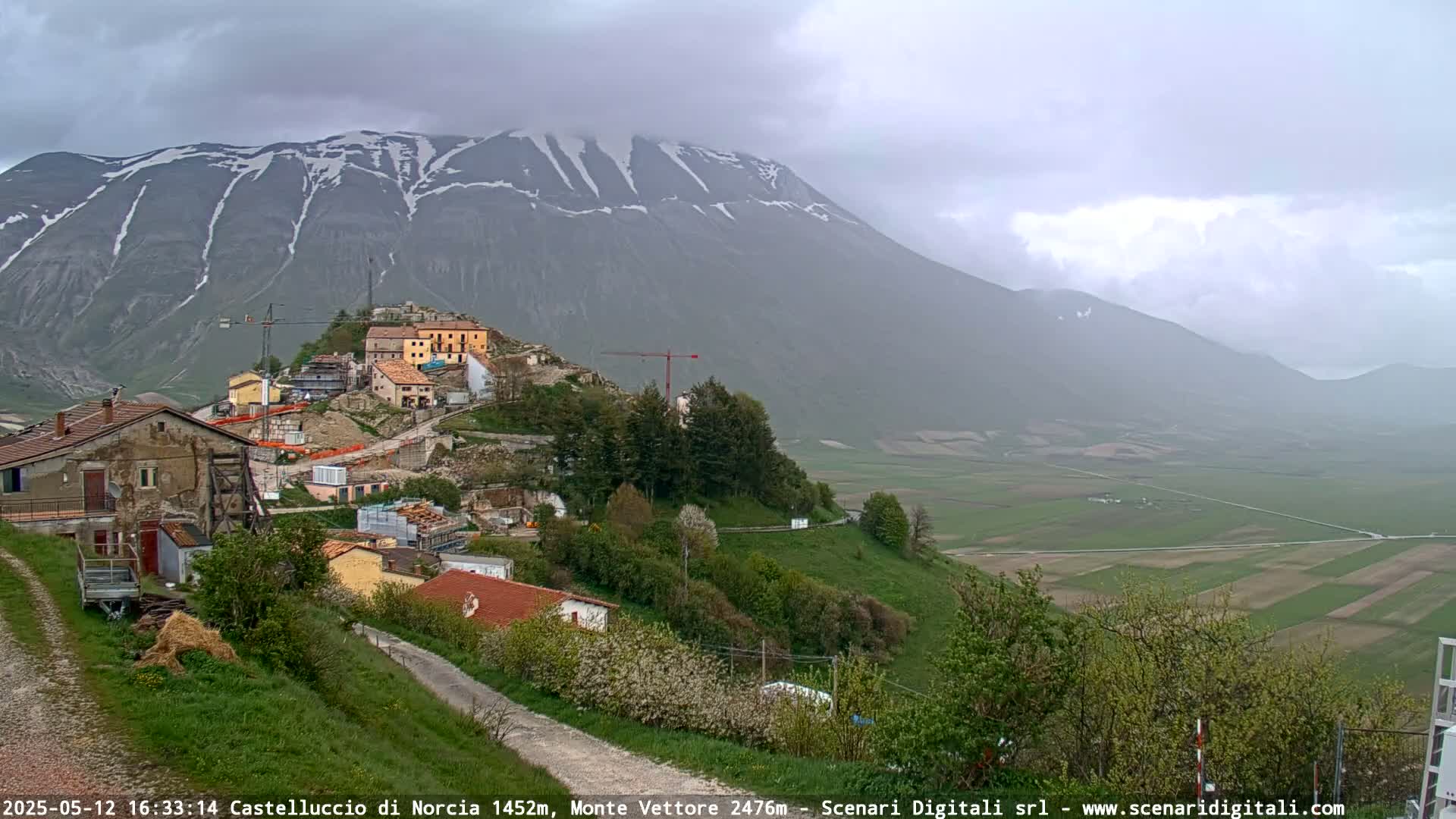 A partially cloudy day reveals a snow-capped mountain overlooking a small village nestled on a hillside, with ongoing construction visible, and a verdant valley stretching into the distance.