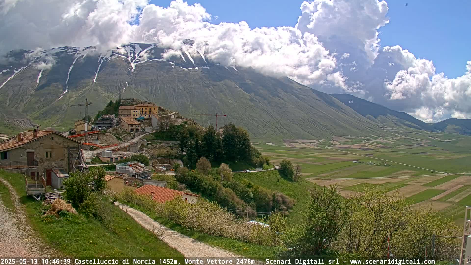 A partly cloudy day reveals a mountainous landscape with snow-capped peaks, a village under construction, and a sprawling green valley below.
