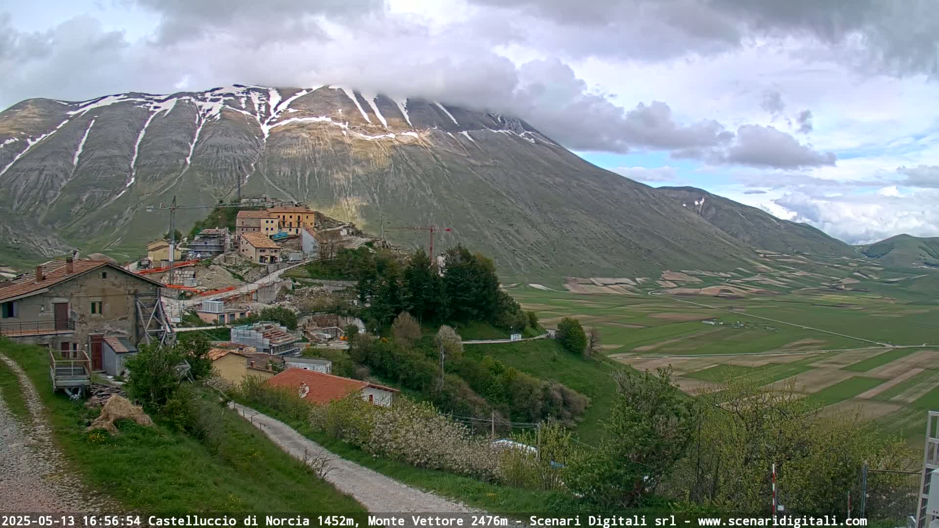 A partially cloudy day reveals a mountain range with snow-capped peaks overlooking a town nestled in a valley of green farmland.