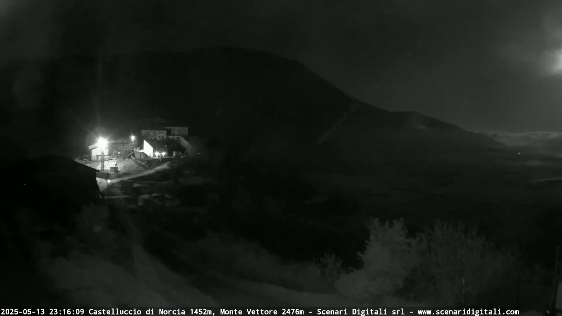 A nighttime, low-light view shows a small village nestled at the base of a dark mountain under a mostly clear sky.