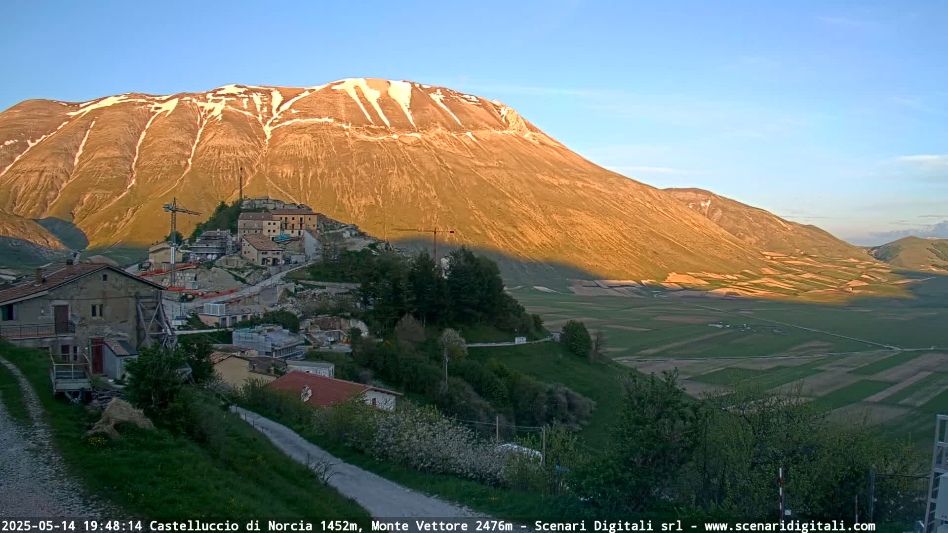 A village nestled in a valley below a large, sunlit mountain with patches of snow, under a clear blue sky.