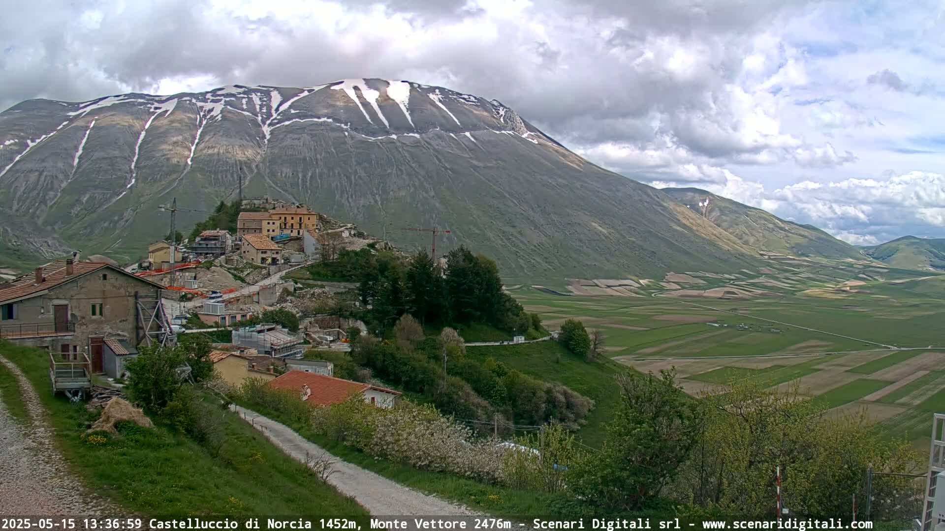 A partly cloudy day reveals a mountain with snow-capped peaks overlooking a village nestled in a valley of green fields.