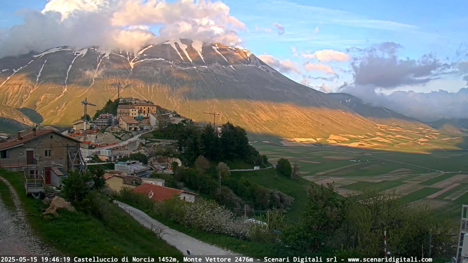 A partially cloudy sunset illuminates a mountain range and a small village nestled below, with fields extending into the valley.