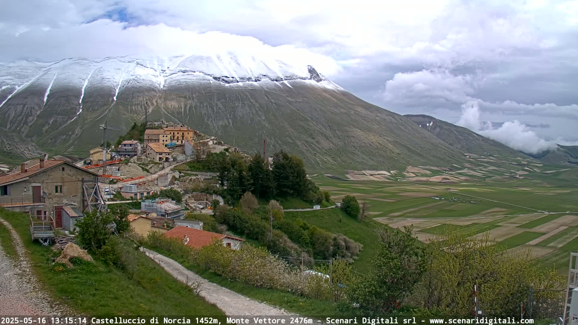 A snow-dusted mountain overlooks a small village nestled in a valley of green farmland under a cloudy sky.