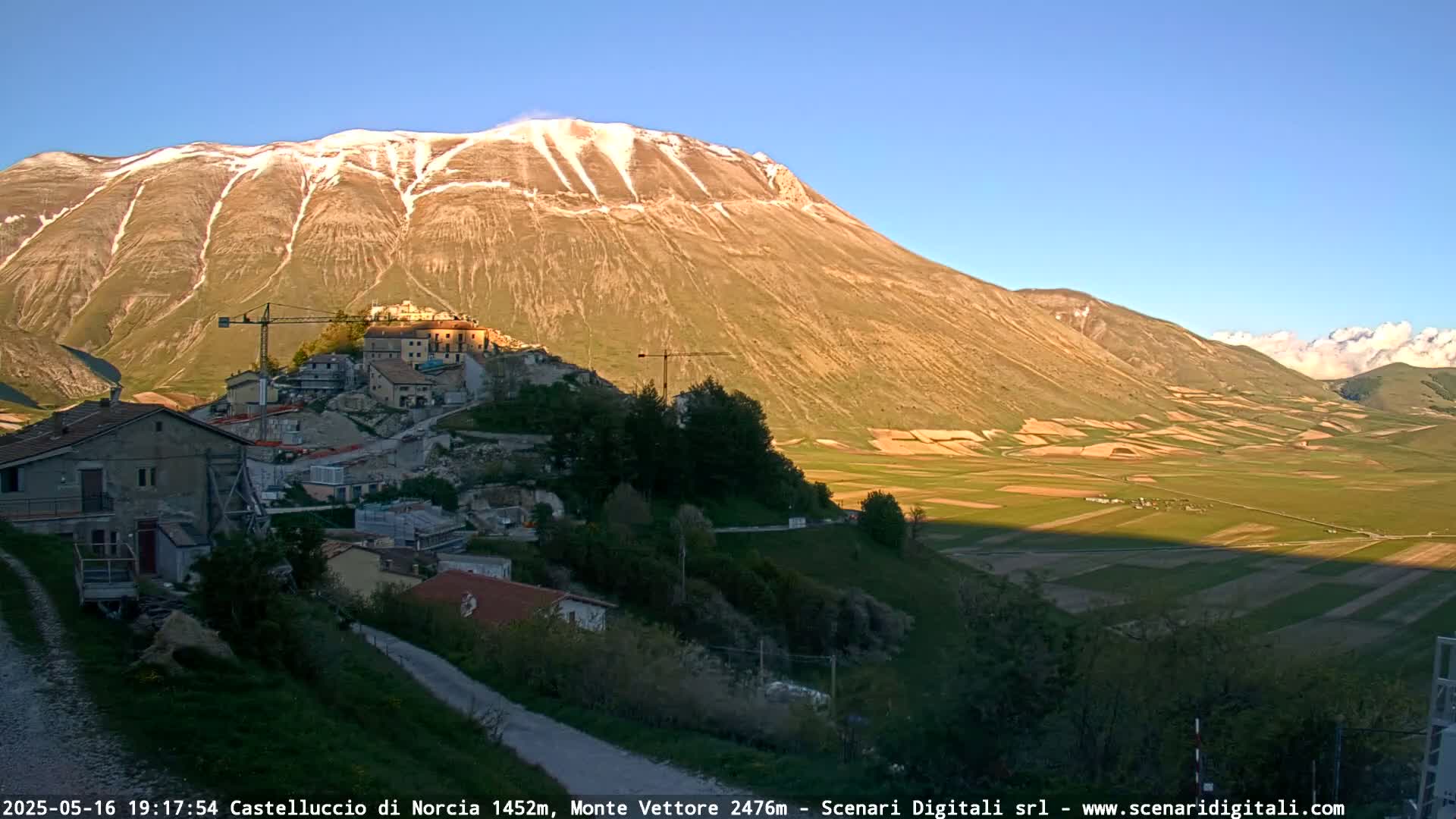 A small village nestled at the base of a large, snow-capped mountain under a clear, sunny sky, overlooks a valley of farmland.