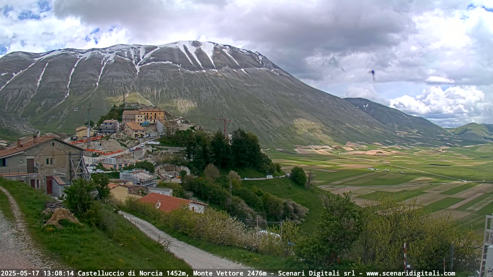A partly cloudy day reveals a mountain range with patches of snow, overlooking a village nestled in a valley of farmland.