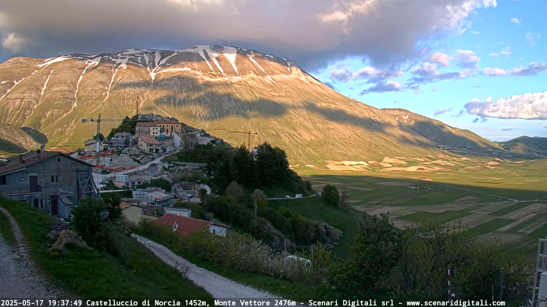 A partly cloudy evening scene shows a village nestled on a hillside below a snow-capped mountain range, with cultivated fields stretching across a valley.