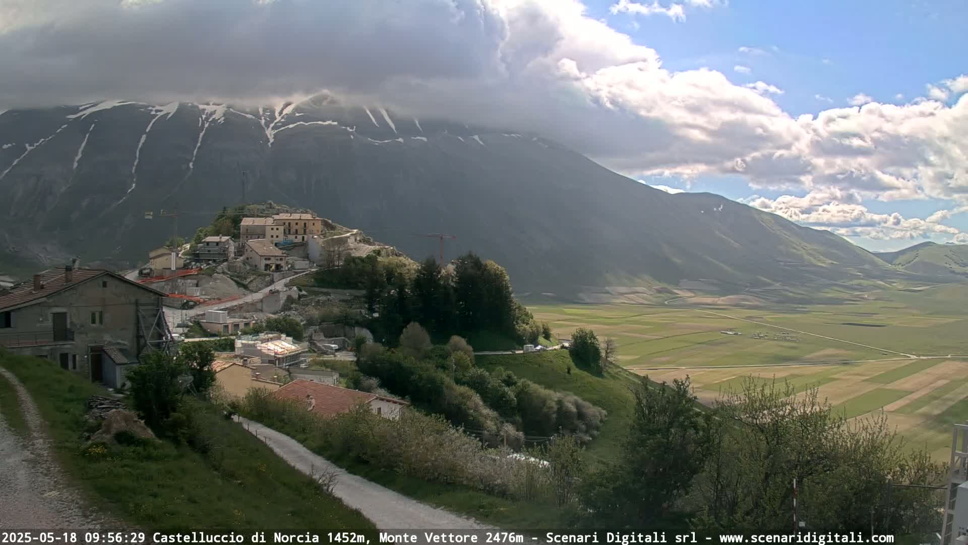 A partially cloudy day reveals a village nestled on a hillside below a snow-capped mountain range and a wide, flat valley of cultivated fields.