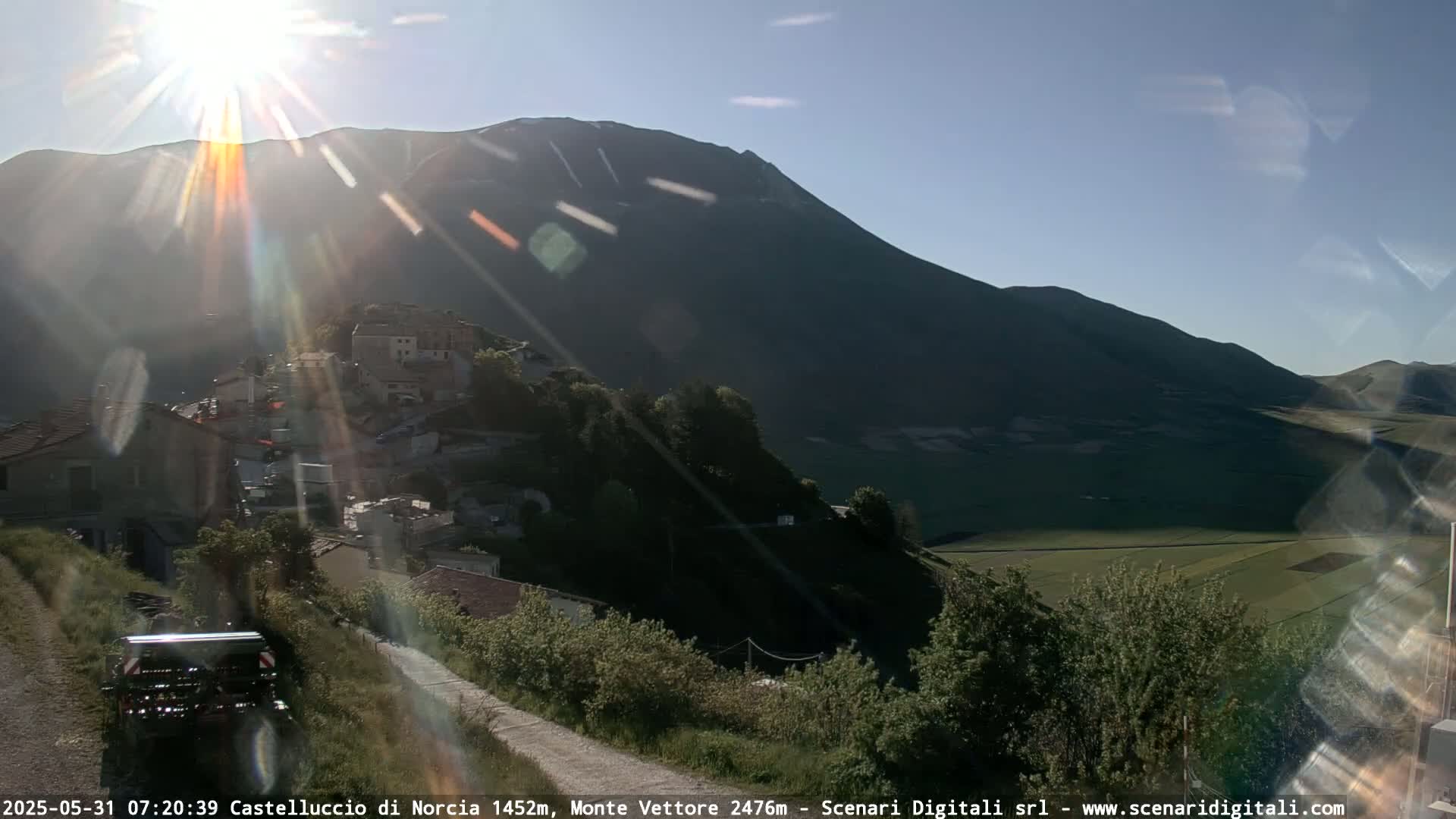 A sun-drenched hillside village nestled in a valley below a large mountain, with a partly sunny sky.