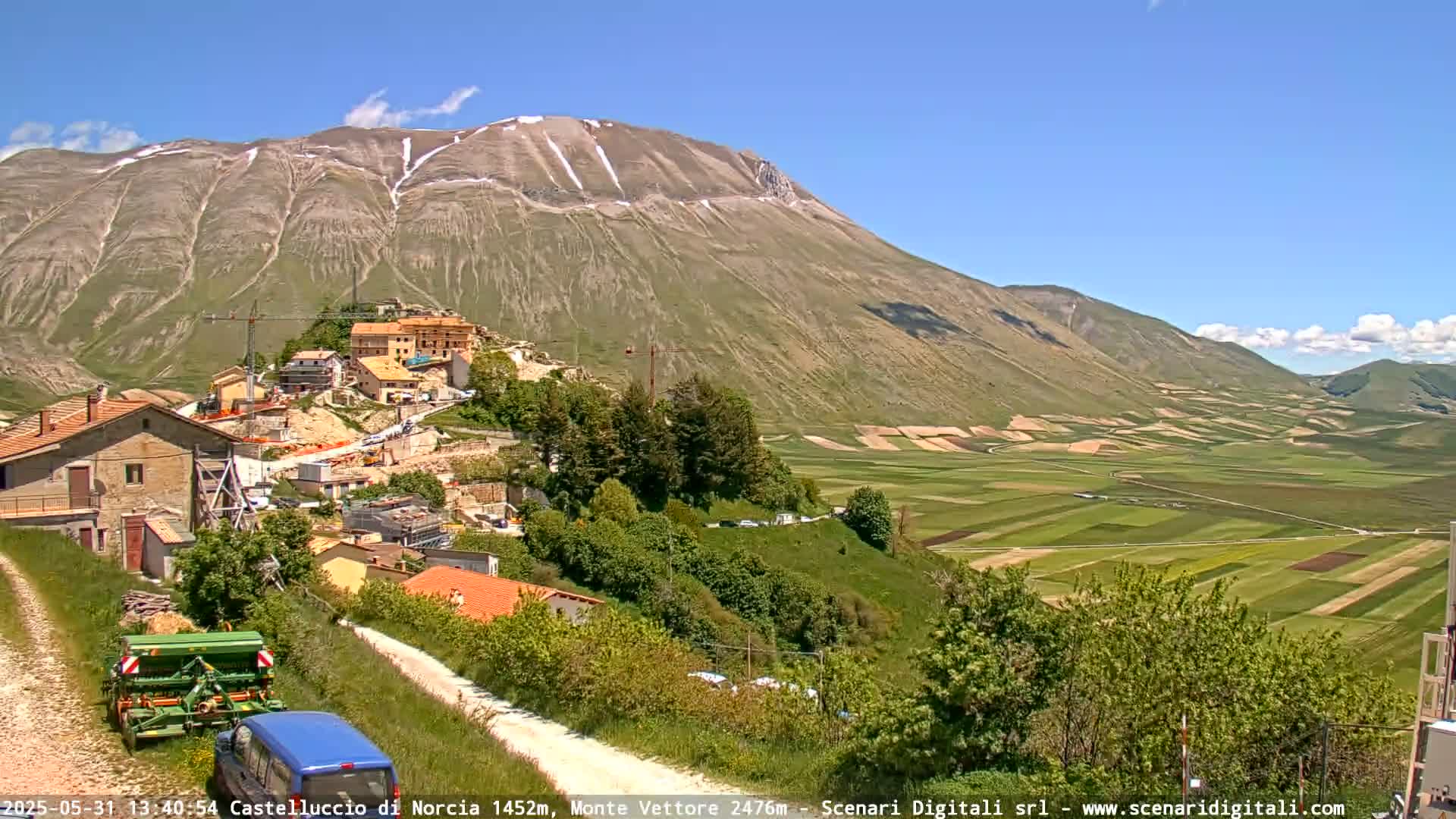 A partly snowy mountain overlooks a village nestled in a valley of verdant farmland under a clear blue sky.