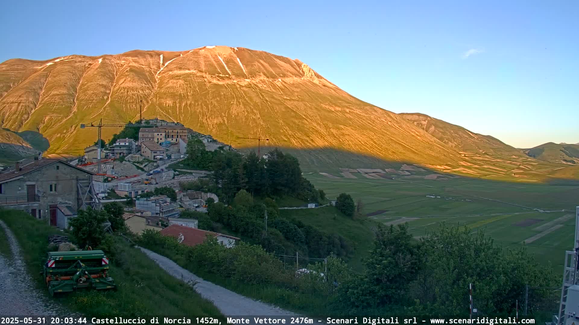 A small village nestled at the base of a large, sunlit mountain, with fields stretching out in the valley below, under a clear evening sky.