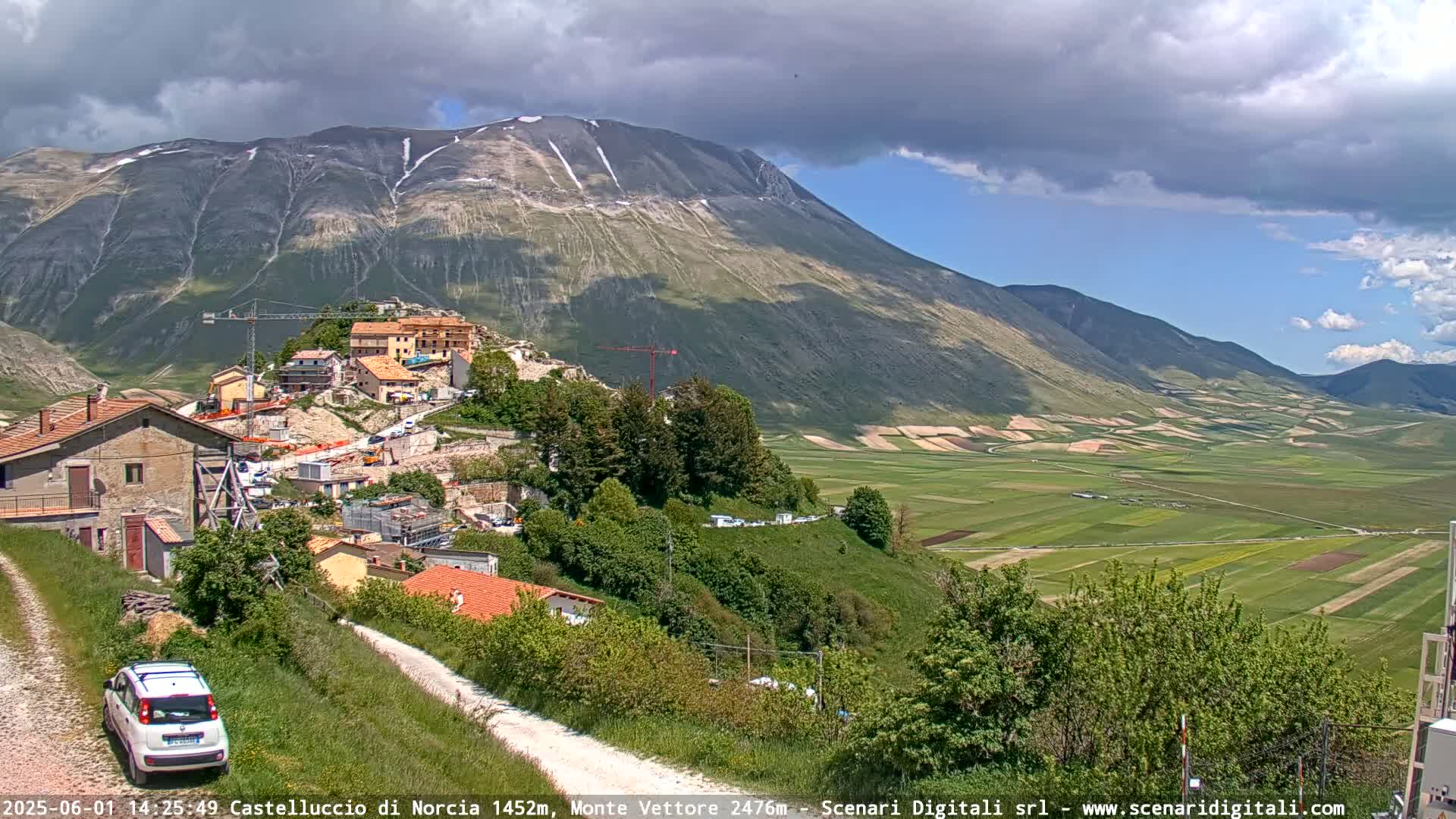 A partly cloudy day reveals a mountain village nestled in a valley next to a large, snow-capped mountain, overlooking a patchwork of green fields.