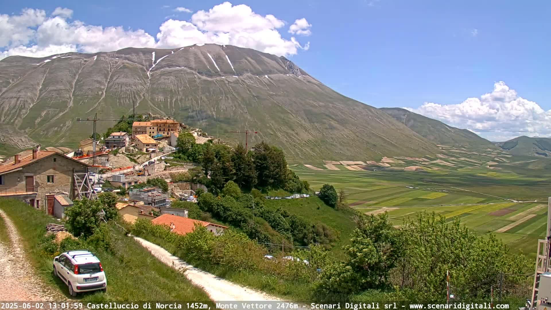 A partly cloudy day reveals a mountainous landscape with a small village nestled amongst green hills and a vast valley of patchwork farmland stretching to the horizon.