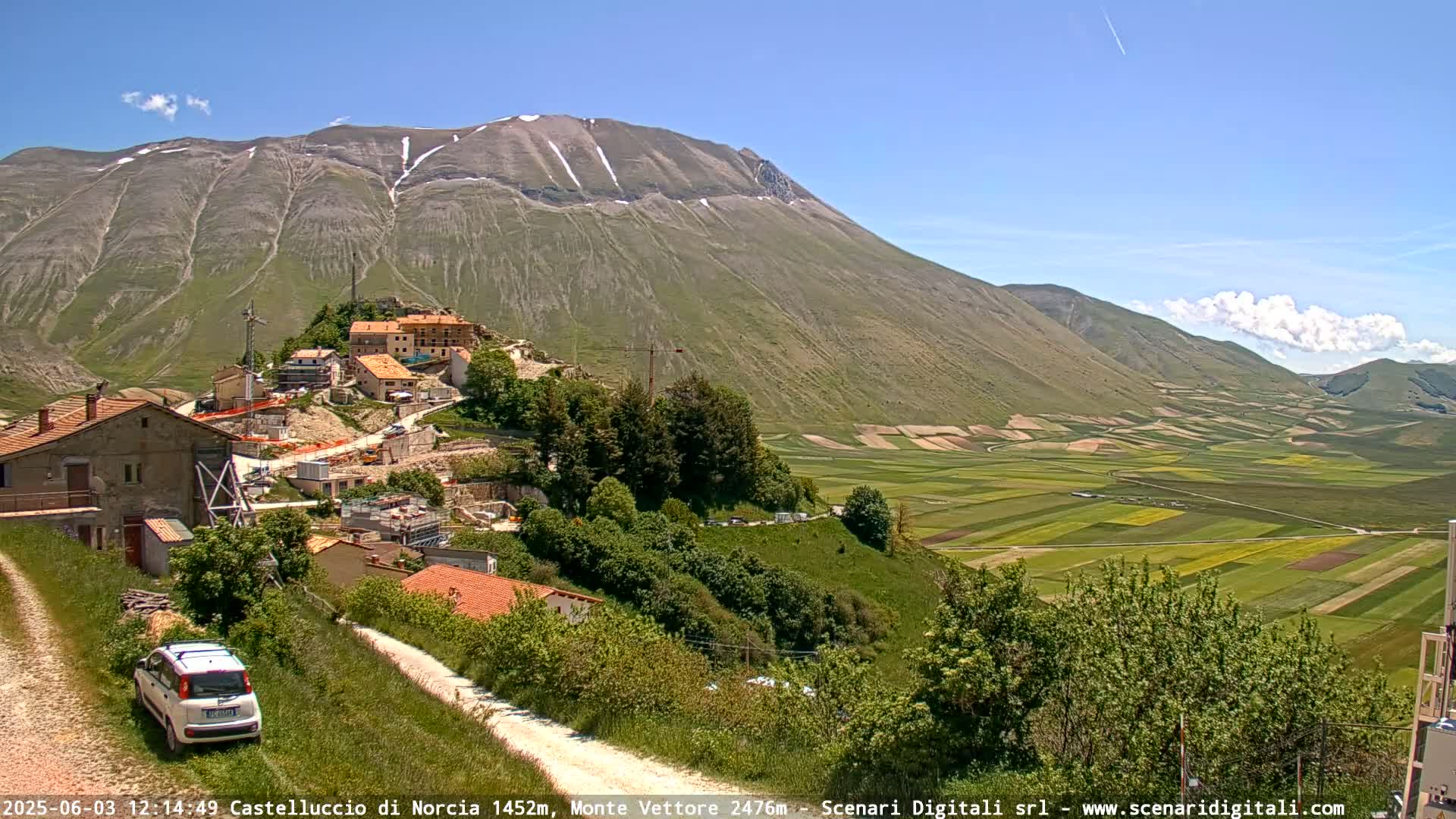 A partly cloudy sunny day shows a small village nestled on a hillside overlooking a vast, patchwork valley of farmland, with a snow-capped mountain in the background.
