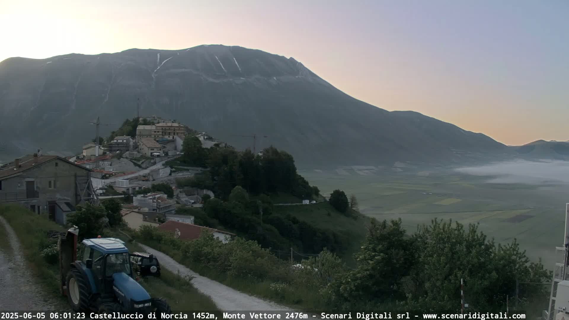 A tractor sits on a dirt road overlooking a hilltop town nestled in a valley below a large mountain, with low-lying fog in the distance under a clear, twilight sky.