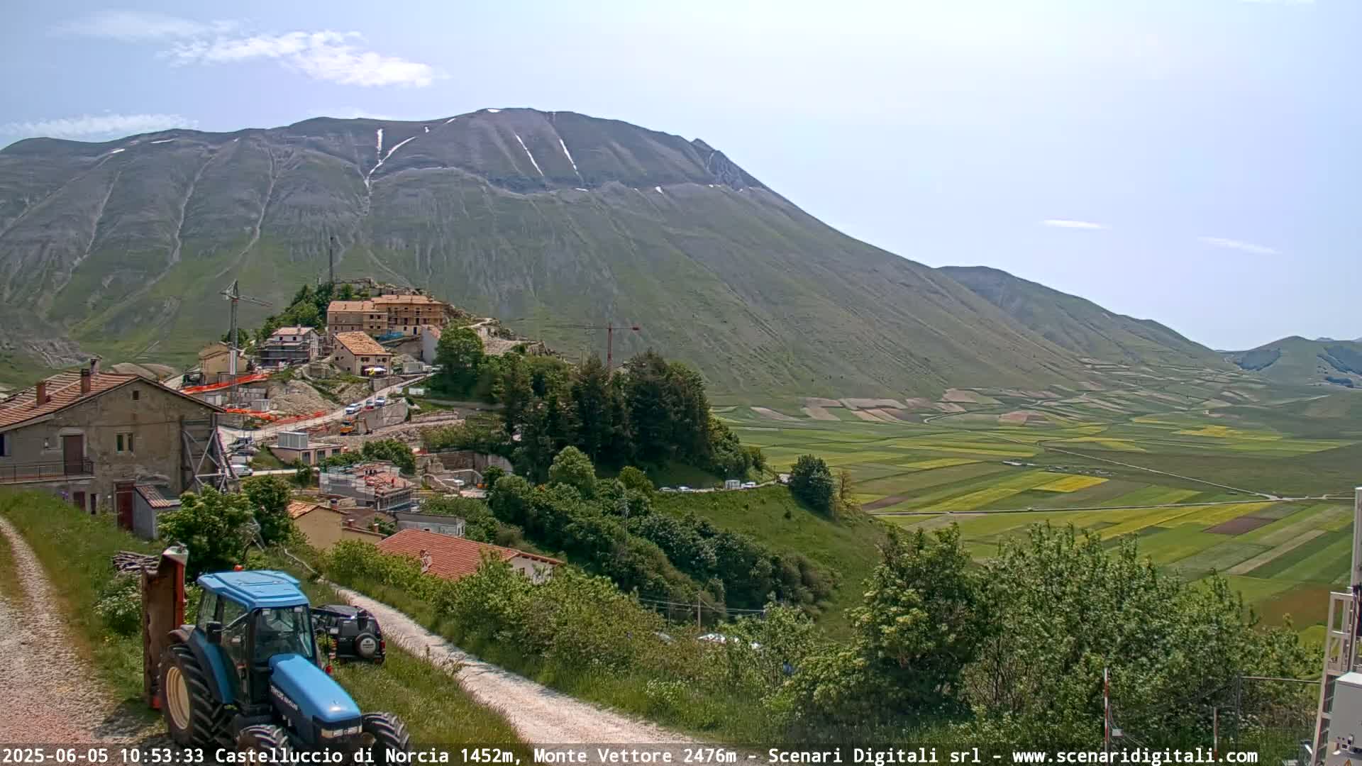 A blue tractor sits on a dirt road overlooking a village nestled against a large mountain with patches of snow, all under a mostly clear sky, with a valley of agricultural fields extending into the distance.