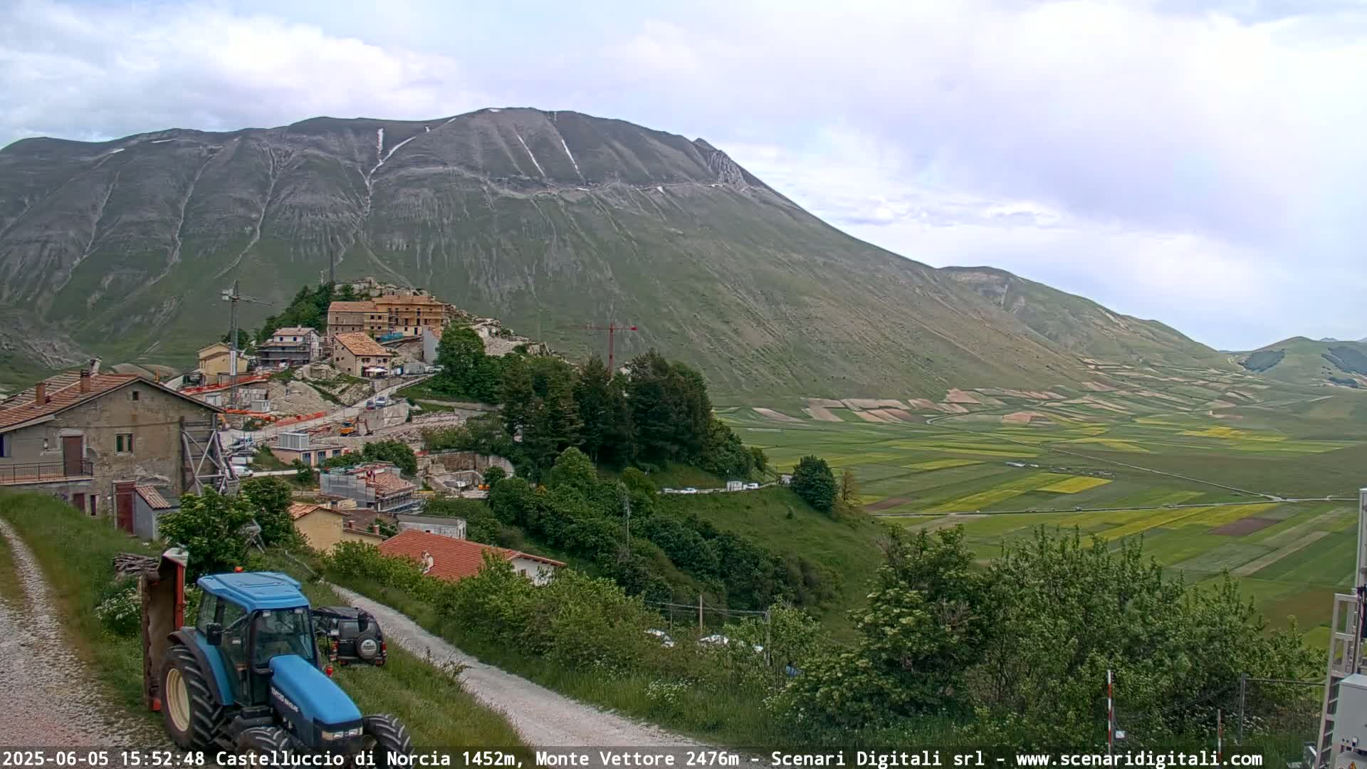 A blue tractor is parked on a dirt road overlooking a village nestled at the base of a large mountain, with patchwork farmlands stretching across a valley under a mostly cloudy sky.