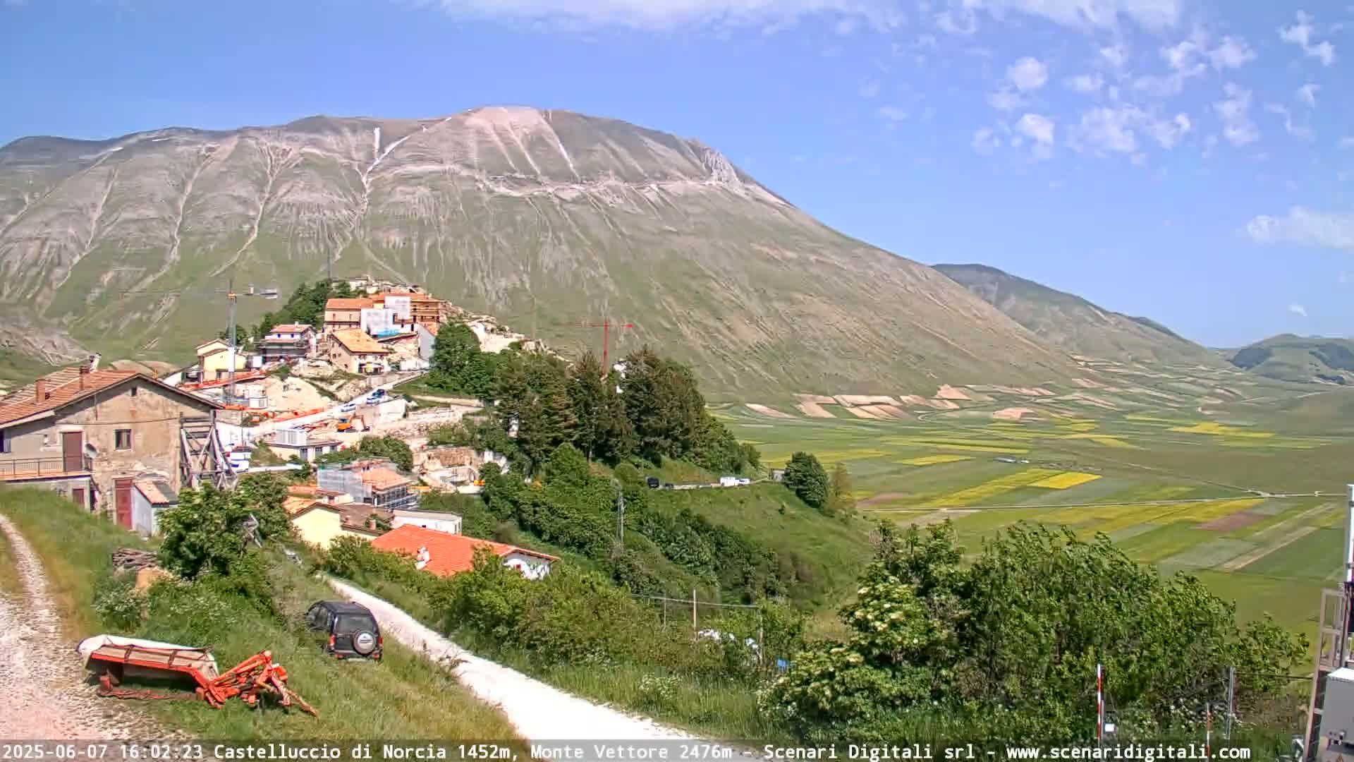 A sunny day reveals a hillside village nestled below a large mountain, overlooking a valley of patchwork farmland.