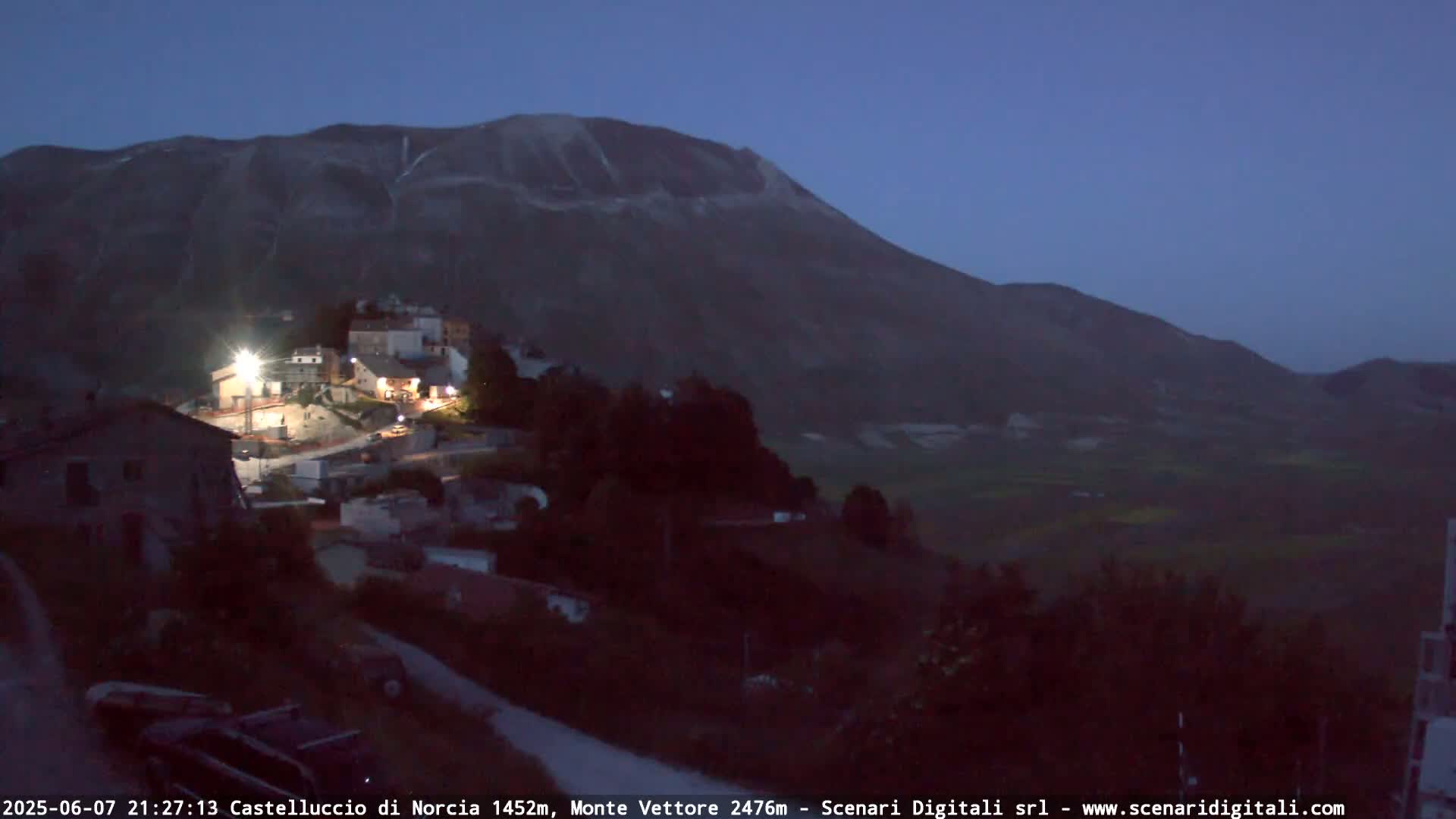 A small, illuminated village nestled at the base of a large, dark mountain under a twilight sky.