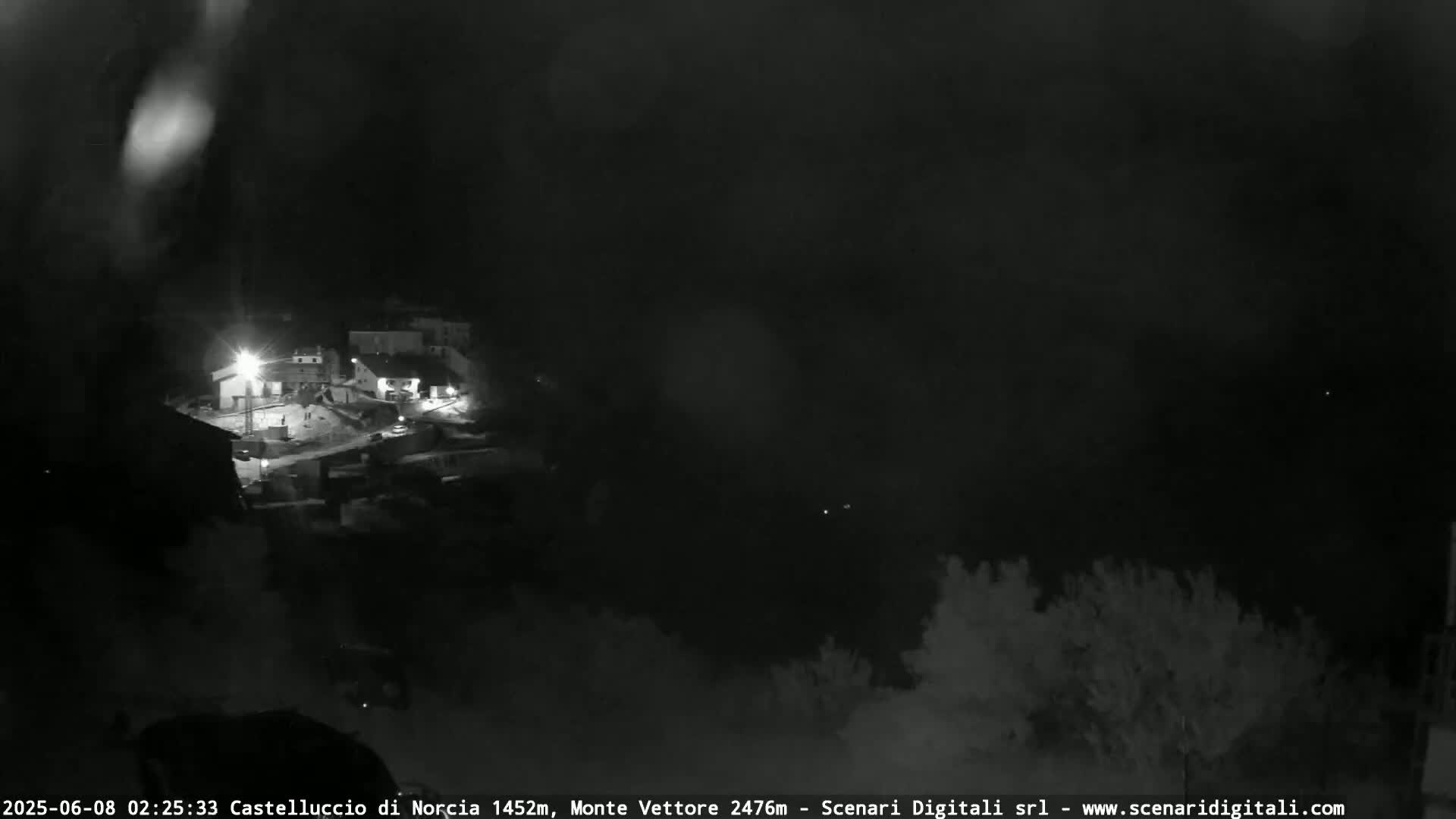 A nighttime, low-light view of a small village nestled in a dark, possibly overcast or foggy landscape.