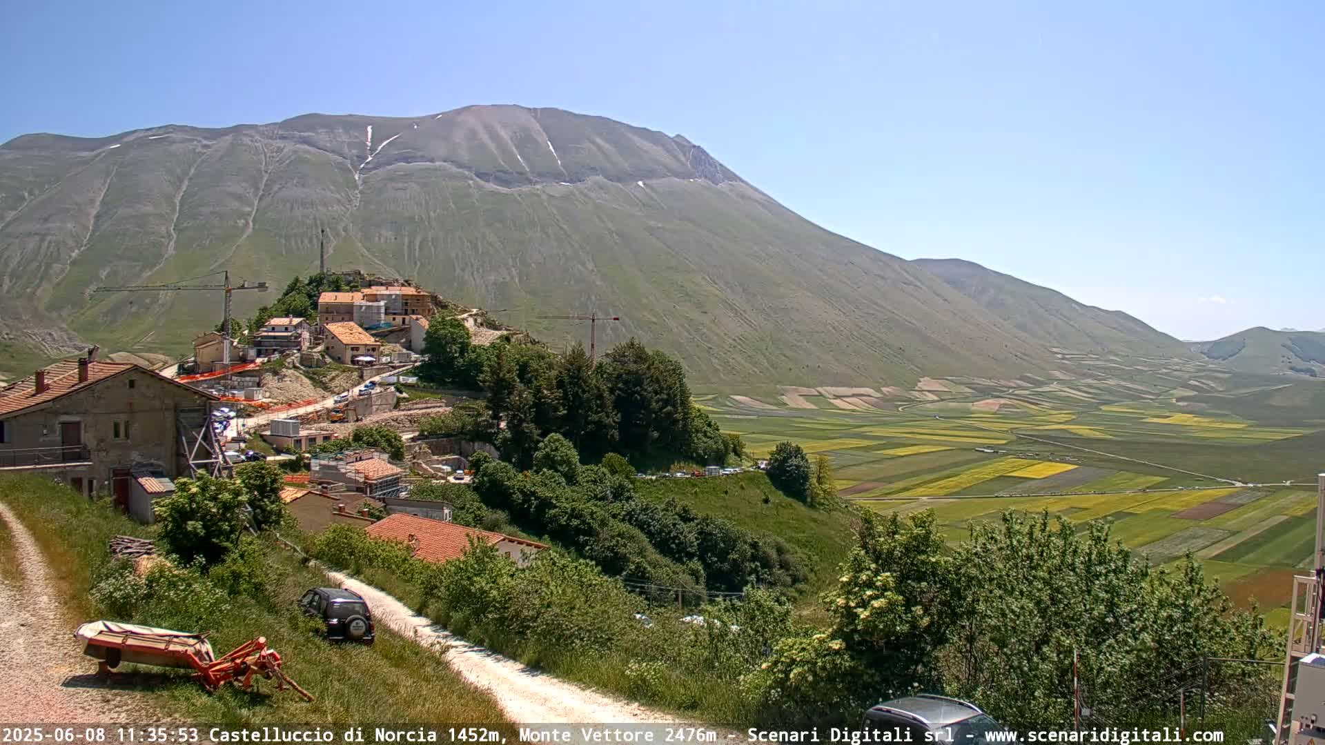A hillside village with construction in progress overlooks a vast valley of patchwork farmland under a clear blue sky.