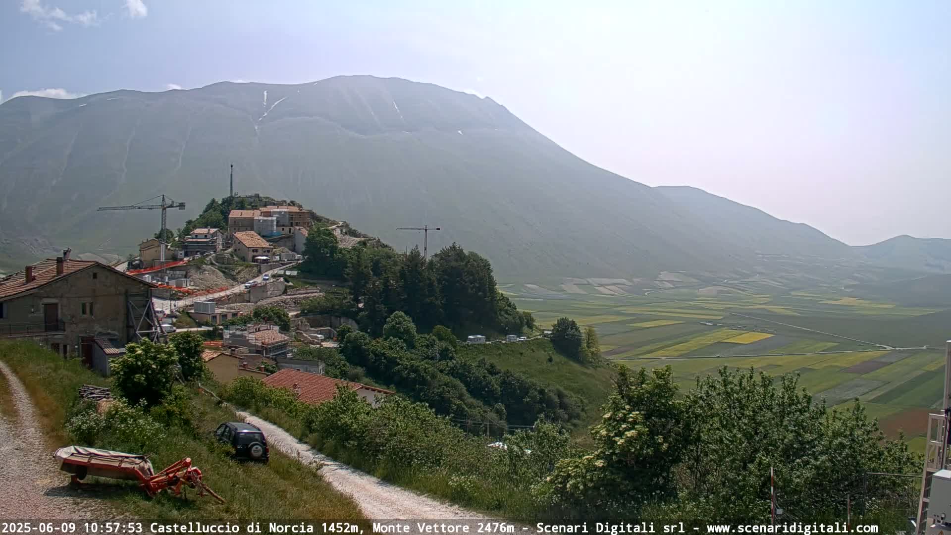 A hazy mountain range overlooks a valley of farmland and a small village with construction cranes, under a mostly clear sky.