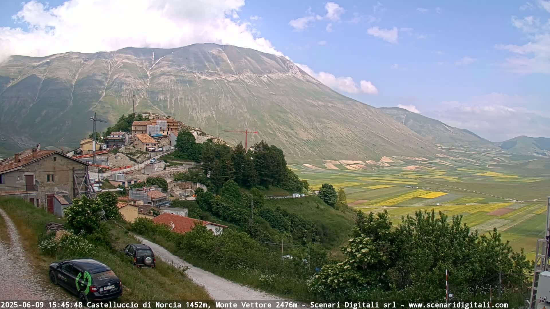 A partly cloudy day reveals a mountain village nestled on a hillside overlooking a vast, patchwork valley of fields, with a large mountain dominating the background.