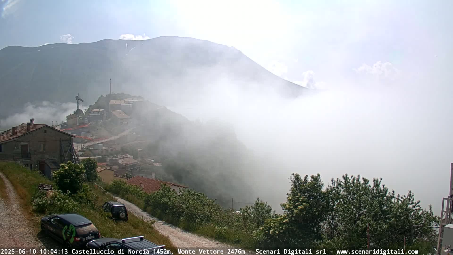 A mountain village is partially obscured by fog, with a mountain range shrouded in low clouds in the background on a partly sunny day.