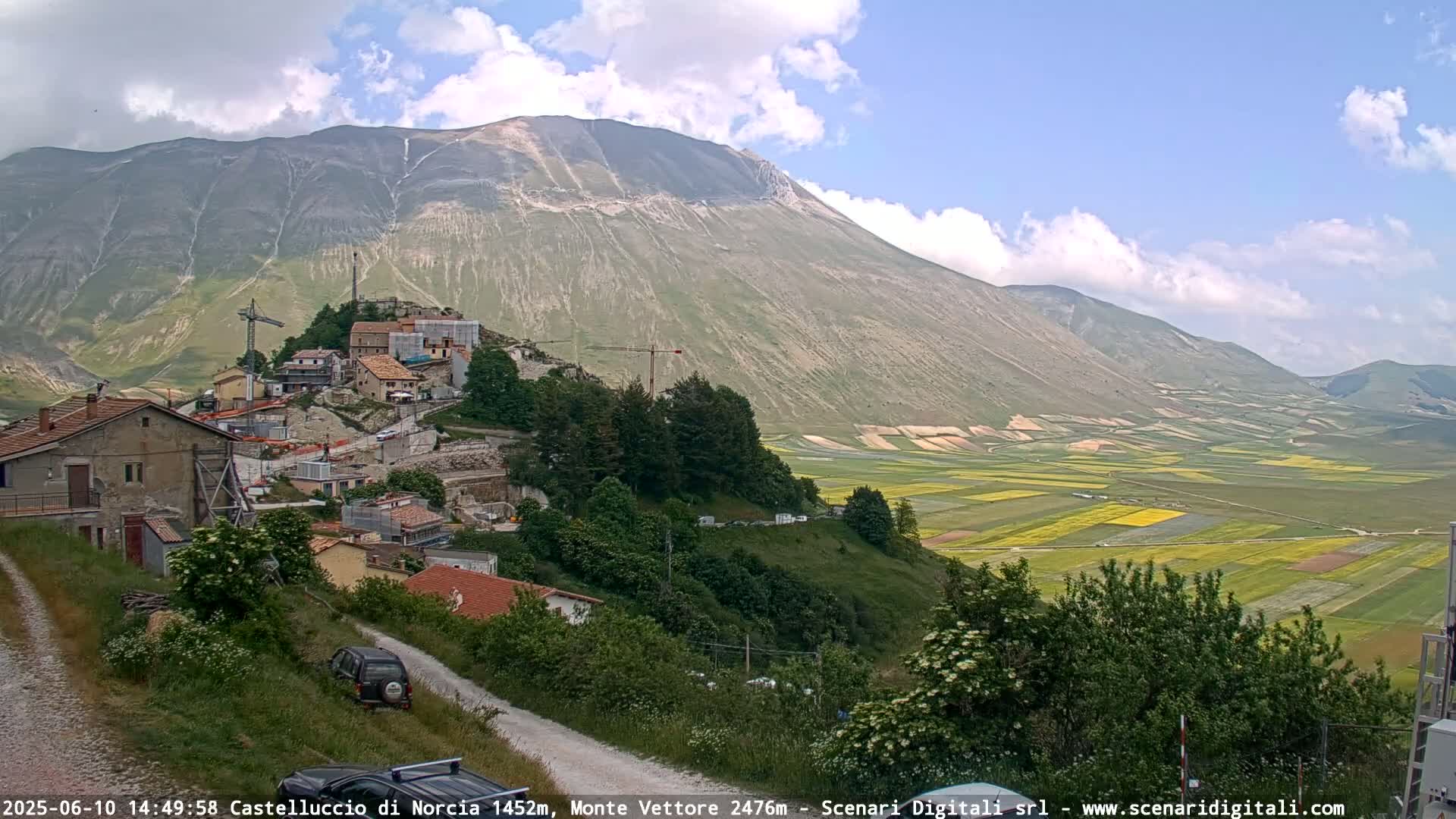 A partly cloudy day reveals a mountain village nestled on a hillside overlooking a vast, patterned valley of farmland.