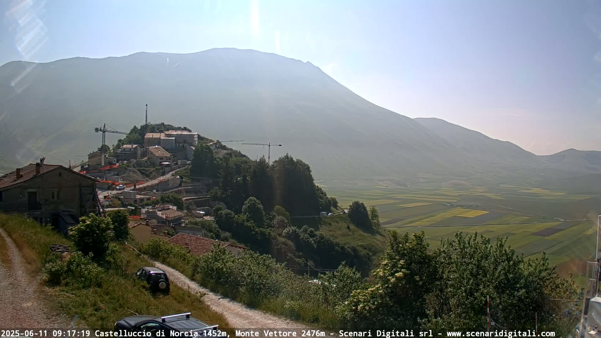 A hillside village with construction cranes is nestled beneath a hazy mountain range overlooking a vast, patchwork farmland under a clear, sunny sky.