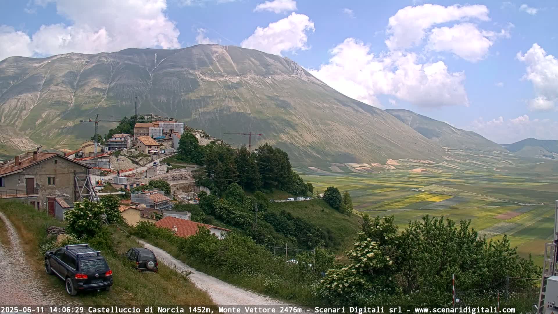 A partly cloudy day reveals a mountain village under construction, nestled on a hillside overlooking a vast, patchwork valley of farmland.
