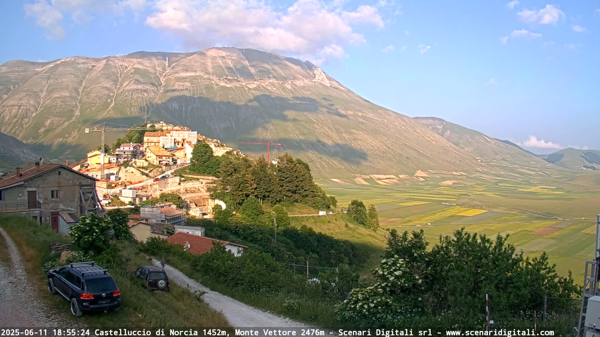 A hillside village nestled against a large mountain, overlooked by a vast, sunlit valley of farmland under a mostly clear sky.