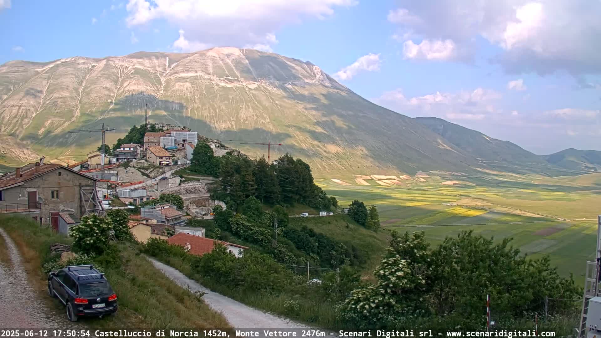 A hilly landscape under a partly cloudy sky shows a village nestled into a mountainside overlooking a vast, flat expanse of farmland.