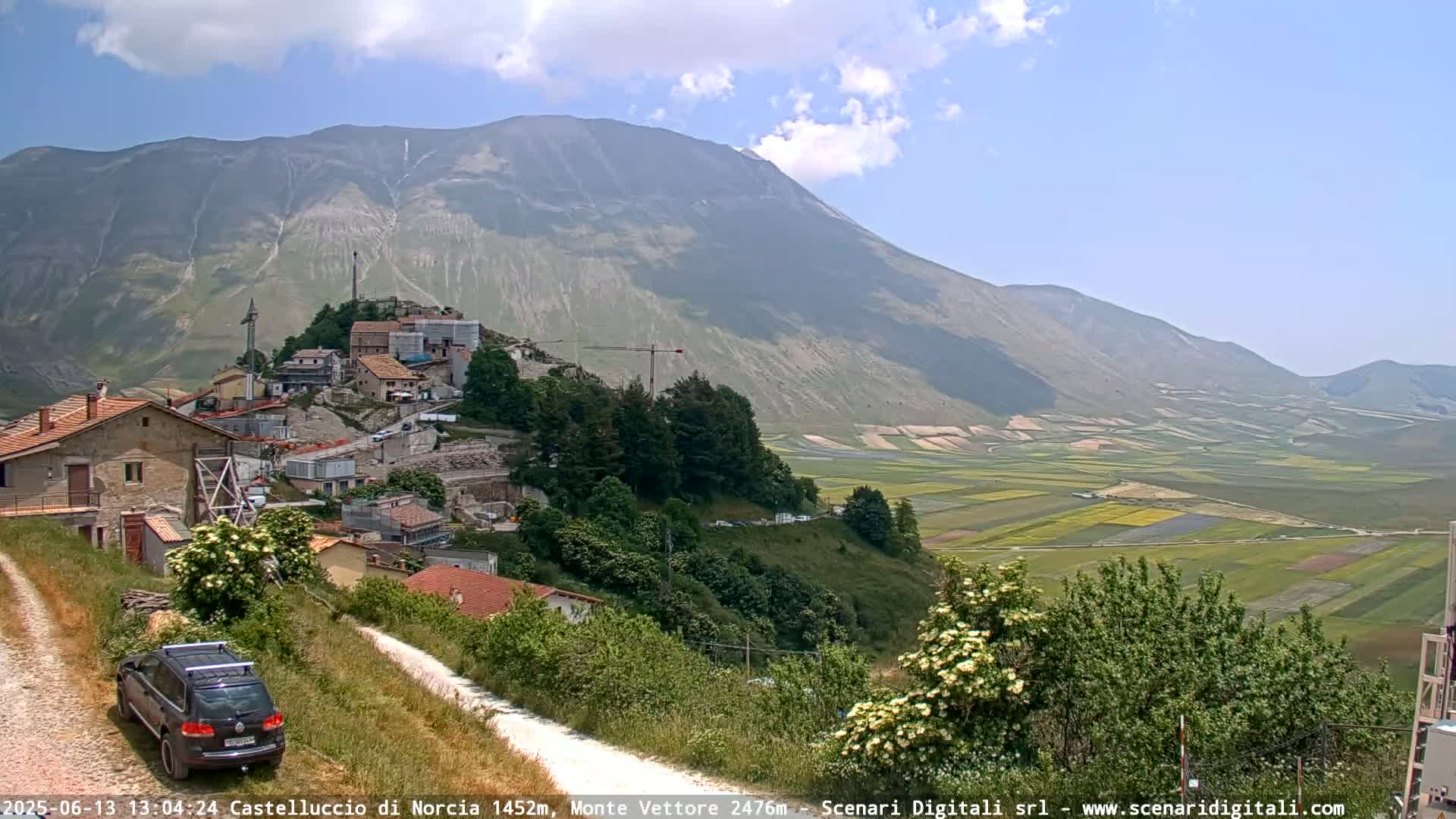 A hilly landscape under a partly cloudy sky shows a village nestled against a mountain, with cultivated fields stretching into the valley below.