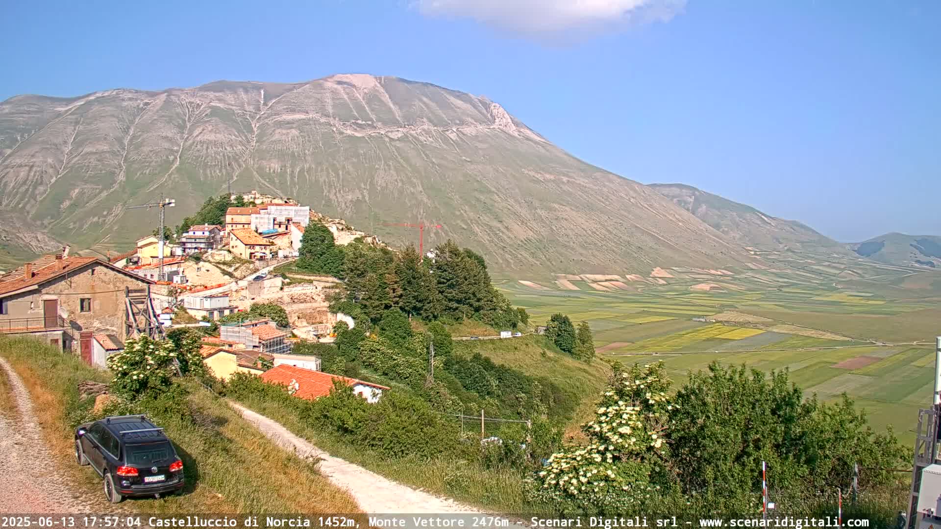 A hillside village overlooks a vast, flat valley of agricultural fields under a clear, sunny sky, with a large mountain in the background.