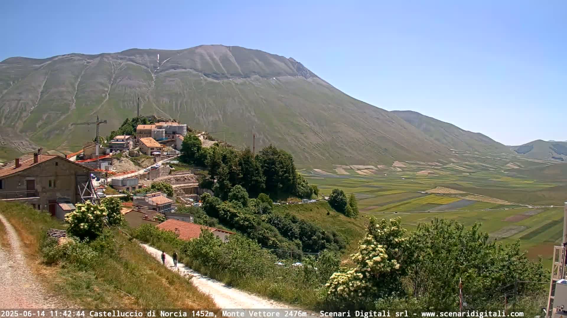A hilltop village with construction equipment is nestled against a large mountain under a clear blue sky, overlooking a vast valley of farmland.