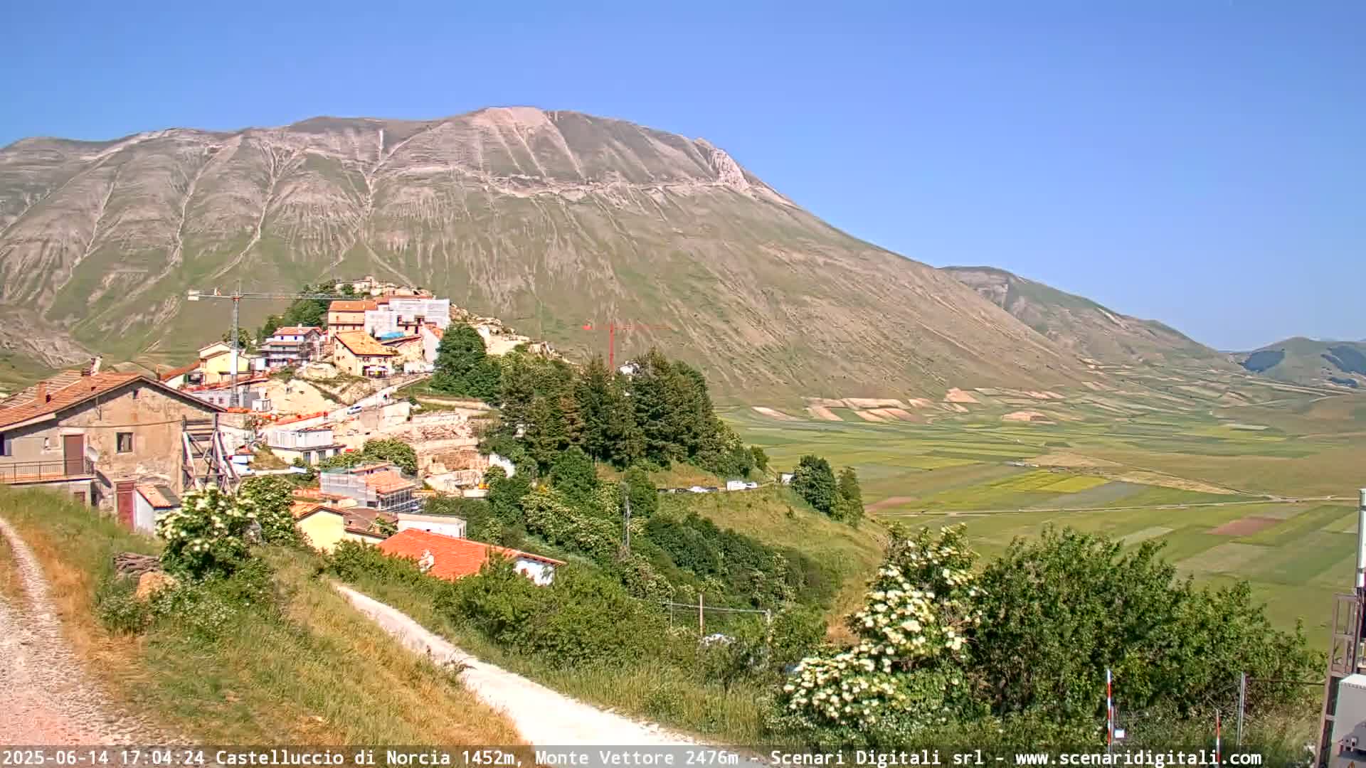 A hilltop village nestled against a large, light brown mountain under a clear blue sky overlooks a valley of patchwork farmlands.