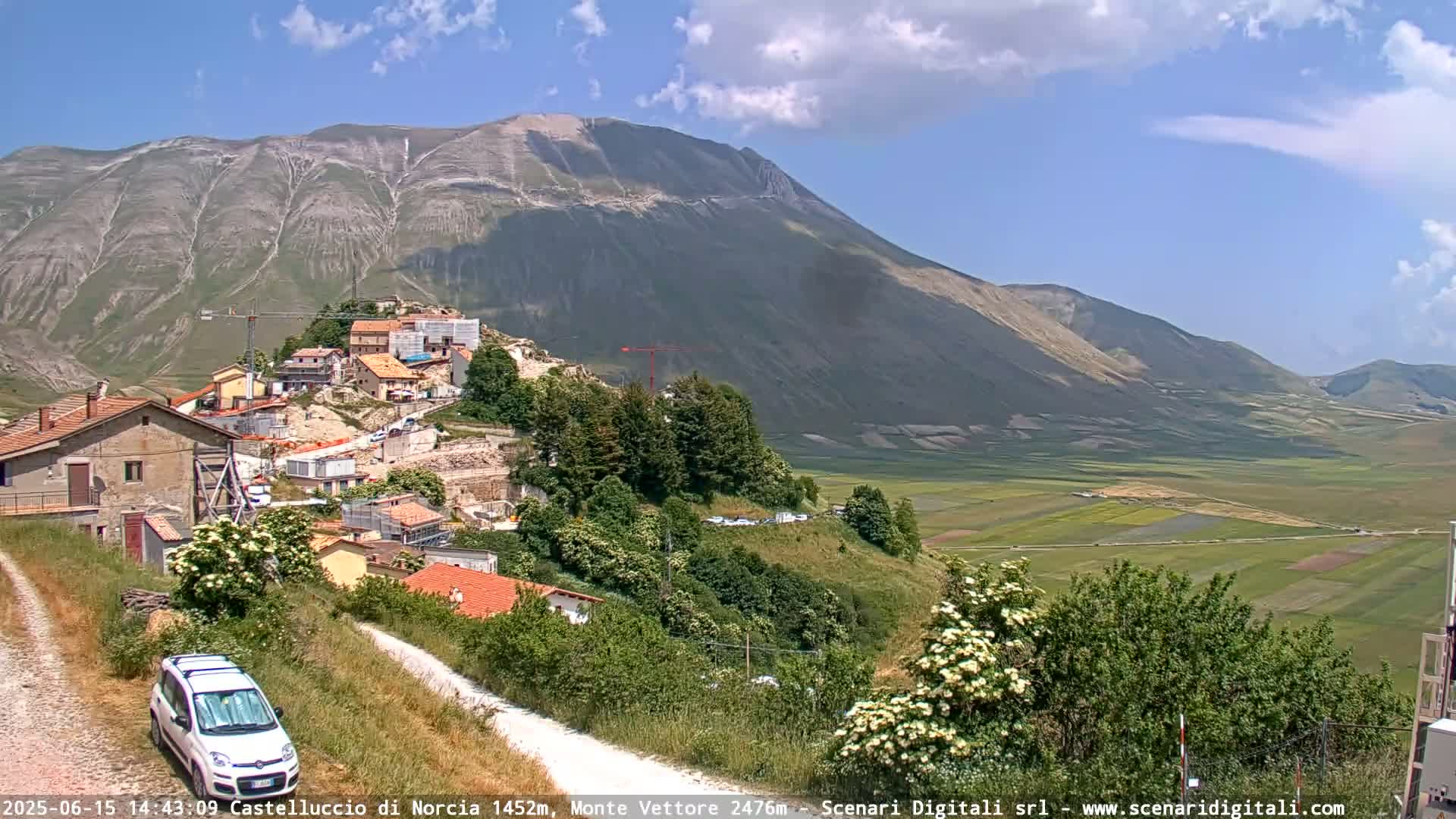 A small village nestled on a hillside beneath a large mountain, with a valley of farmland visible in the distance under a mostly sunny sky.
