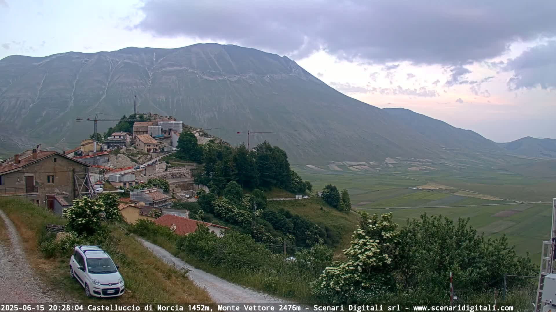 A village nestled in a valley below a large mountain under a partly cloudy sky, with construction visible in the village and a car parked on a nearby road.