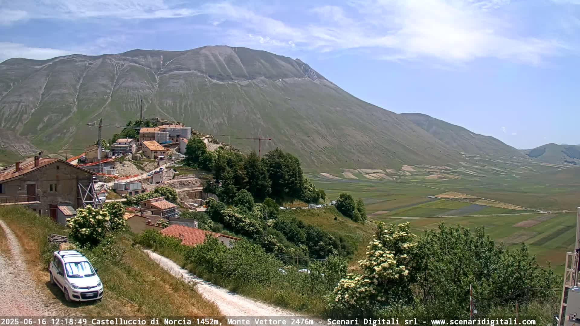 A small village nestled on a hillside overlooking a vast, flat green valley under a mostly sunny sky.